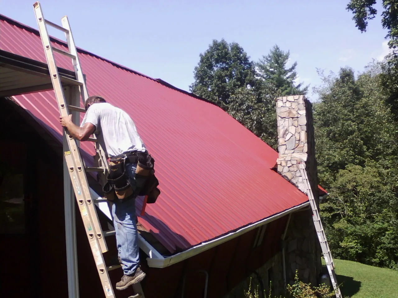 Roofer on a ladder beside a red metal roof and stone chimney — Soffit & Fascia Repair by Roofing Brothers in Stayton